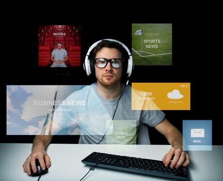 technology, communication, mass media, virtual reality and people concept - young man in headset and eyeglasses with pc computer keyboard over virtual screensの写真素材