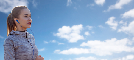 fitness, sport, people, technology and lifestyle concept - happy woman running and listening to music in earphones over blue sky and clouds backgroundの写真素材