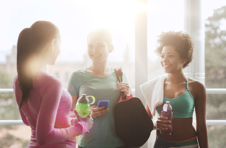 fitness, sport, training and lifestyle concept - group of happy women with bottles of water, smartphone and bag talking in gymの写真素材