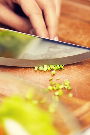 healthy eating, cooking, vegetarian food, kitchenware and people concept - close up of woman chopping green onion with knife on wooden cutting boardの写真素材