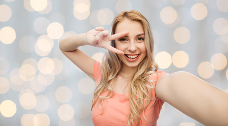 emotions, expressions and people concept - happy smiling young woman taking selfie and showing peace hand sign over holidays lights backgroundの写真素材