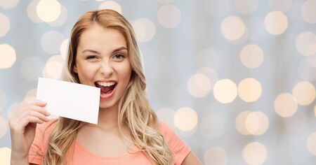advertisement, invitation, message and people concept - smiling young woman or teenage girl with blank white paper card over holidays lights backgroundの写真素材