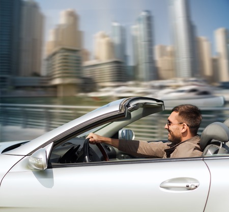 auto business, transport, leisure and people concept - happy man driving cabriolet car over dubai city port backgroundの写真素材
