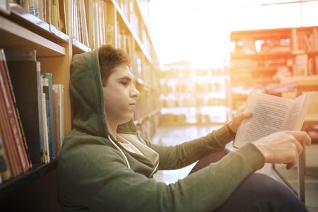 people, knowledge, education, literature and school concept - student boy or young man sitting on floor reading book in libraryの写真素材
