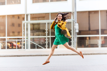 technology, communication and people concept - smiling young woman or girl calling on smartphone on city street and jumping highの写真素材