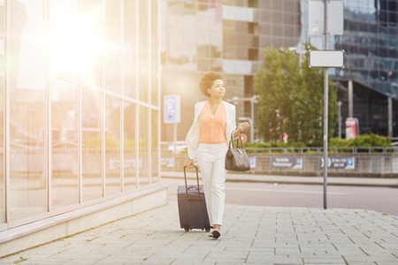 travel, business trip, people and tourism concept - young african american  woman with travel bag walking down city streetの写真素材