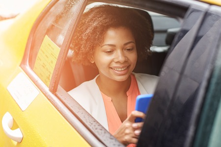business trip, transportation, travel, gesture and people concept - young smiling african american woman texing on smartphone in taxi at city streetの写真素材