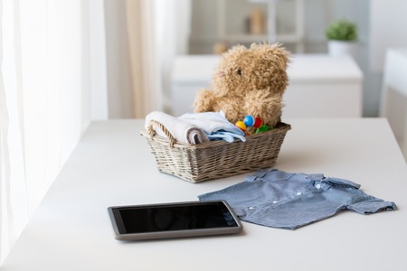 babyhood, motherhood, clothing, technology and object concept - close up of baby clothes and toys for newborn boy in basket with tablet pc computer at homeの写真素材