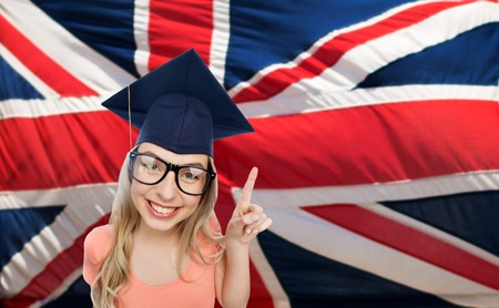 people, graduation and national education concept - smiling young student woman in mortarboard and eyeglasses pointing finger up over english flagの写真素材