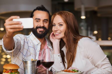 leisure, technology, date, people and holidays concept - happy couple having dinner and taking selfie by smartphone at restaurantの写真素材
