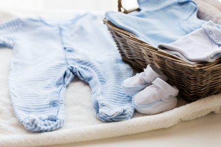 clothing, babyhood, motherhood and object concept - close up of white baby bootees with pile of clothes and towel for newborn boy in basket on tableの写真素材