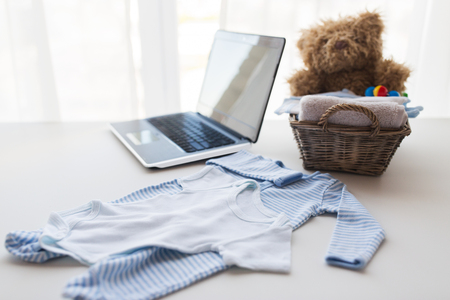 babyhood, motherhood, clothing, technology and object concept - close up of baby clothes and toys for newborn boy in basket with laptop computer at homeの写真素材