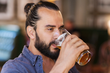 people, drinks, alcohol and leisure concept - happy young man drinking beer from glass at bar or pubの写真素材