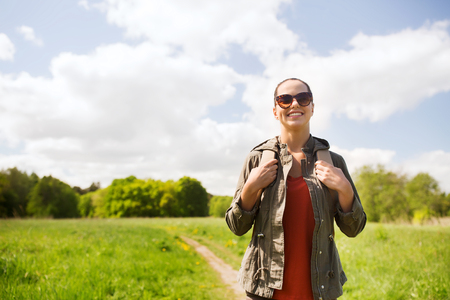 travel, hiking, backpacking, tourism and people concept - happy young woman in sunglasses with backpack walking along country road outdoorsの写真素材