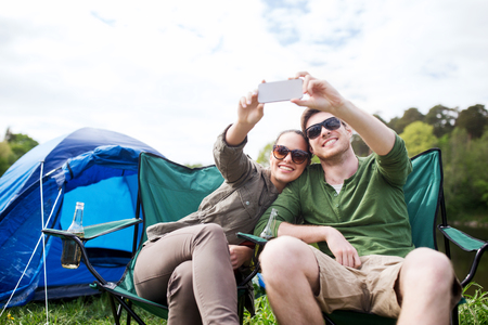 travel, hiking, technology, tourism and people concept - smiling couple of travelers taking selfie by smartphone at campingの写真素材