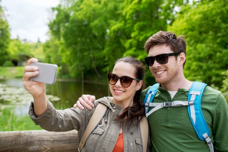 travel, hiking, backpacking, tourism and people concept - smiling couple with backpacks taking selfie by smartphone in natureの写真素材