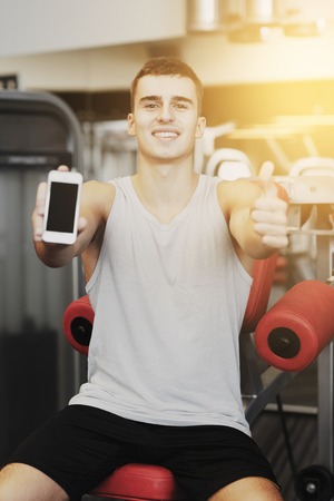 sport, bodybuilding, lifestyle, technology and people concept - smiling young man showing smartphone and thumbs up gesture in gymの写真素材