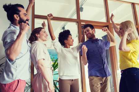 business, triumph, gesture, people and teamwork concept - happy creative team raising hands up and celebrating victory in officeの写真素材
