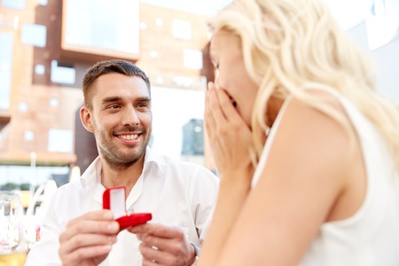 love, anniversary, surprise, people and holidays concept - happy man with engagement ring making proposal to woman at restaurantの写真素材