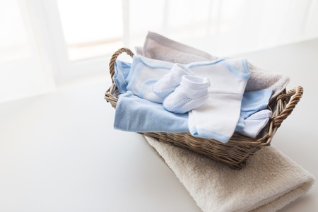 clothing, babyhood, motherhood and object concept - close up of white baby bootees with pile of clothes and towel for newborn boy in basket on tableの写真素材