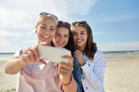 summer vacation, holidays, travel, technology and people concept- group of smiling young women taking sulfide with smartphone on beachの写真素材