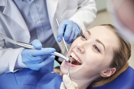 people, medicine, stomatology and health care concept - close up of dentists and assistant with mirror, drill and dental air water gun spray treating female patient teeth at dental clinicの写真素材