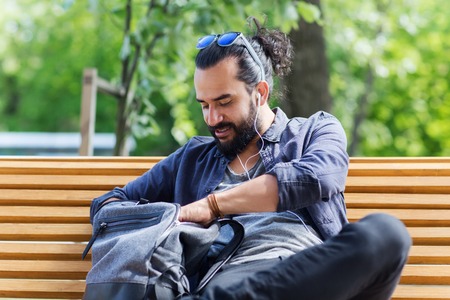 travel, tourism, lifestyle and people concept - man with earphones and sunglasses sitting on city bench and looking for something in his backpackの写真素材
