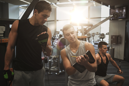 sport, fitness, lifestyle and people concept - men with clipboard taking notes and exercising on gym machineの写真素材