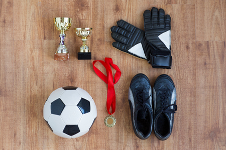 sport, achievement, championship and success concept - close up of soccer ball, football boots and goalkeeper gloves with golden medal and cups on wooden backgroundの写真素材
