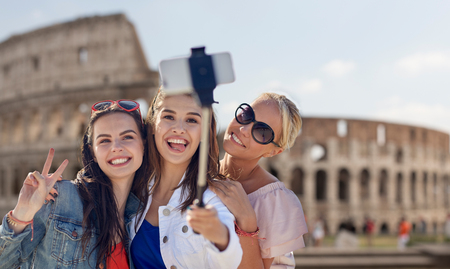 summer vacation, holidays, travel, technology and people concept- group of smiling young women taking picture with smartphone on selfie stick over coliseum in rome backgroundの写真素材