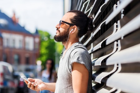people, music, technology, leisure and lifestyle - happy young hipster man with earphones and smartphone on city streetの写真素材
