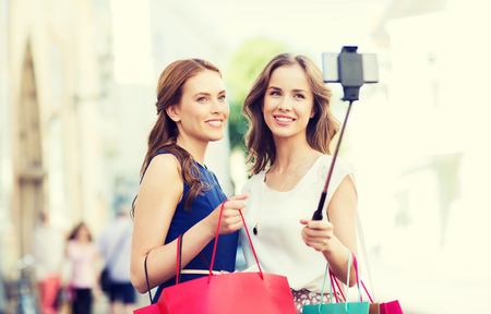 sale, consumerism, technology and people concept - happy young women with shopping bags and smartphone selfie stick taking picture on city streetの写真素材
