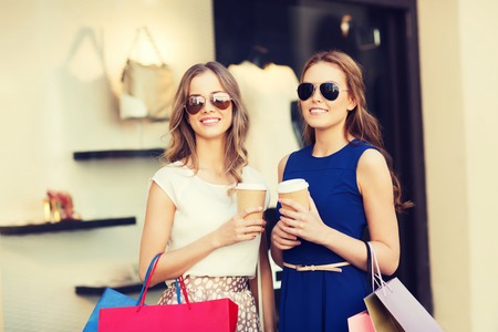 sale, consumerism and people concept - happy young women with shopping bags and coffee paper cups at shop window in cityの写真素材