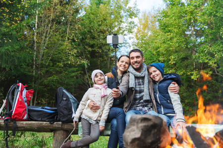 camping, travel, tourism, hike and people concept - happy family sitting on bench and taking picture with smartphone on selfie stick at campfire in woodsの写真素材