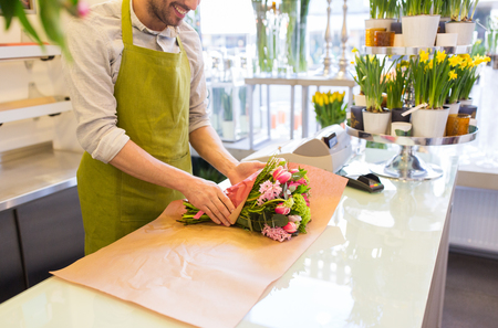 people, shopping, sale, floristry and consumerism concept - close up of happy florist man wrapping flowers in paper at flower shopの写真素材
