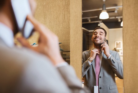 sale, shopping, fashion, style and people concept - happy young man or businessman trying suit and tie on and calling on smartphone at clothing store mirrorの写真素材