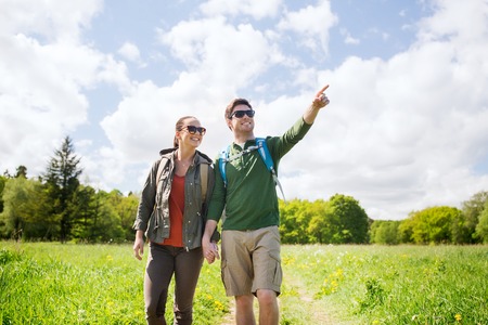 travel, hiking, backpacking, tourism and people concept - happy couple with backpacks walking along country road outdoors and pointing finger to somethingの写真素材
