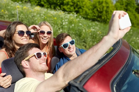 leisure, road trip, travel and people concept - happy friends driving in cabriolet taking selfie by...