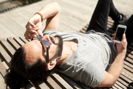 people, music, technology, leisure and lifestyle - happy young hipster with earphones and smartphone...
