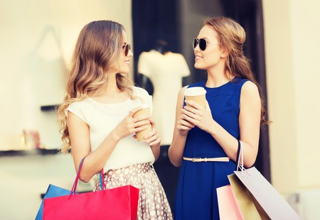 sale, consumerism and people concept - happy young women with shopping bags and coffee paper cups talking at shop window in cityの写真素材