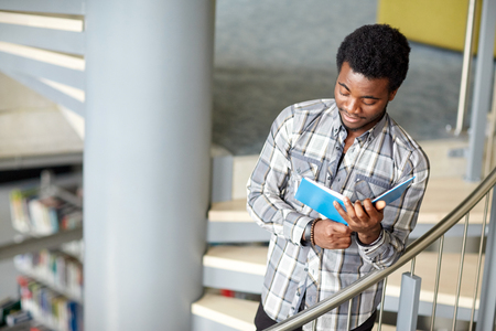 education, high school, university, learning and people concept - happy african american student boy or young man reading book on stairs at libraryの写真素材