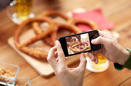 people, food, and technology concept - close up of hands with smartphone picturing beer and pretzel at bar or pubの写真素材