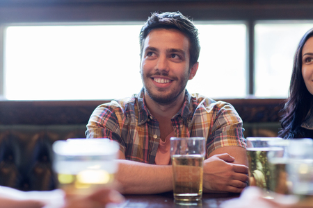 people, leisure, friendship and communication concept - happy man with friends drinking beer and watching sport game or football match at bar or pubの写真素材