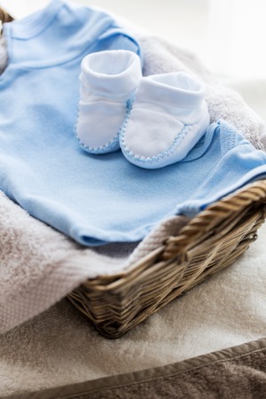 clothing, babyhood, motherhood and object concept - close up of white baby bootees with pile of clothes and towel for newborn boy in basket on tableの写真素材