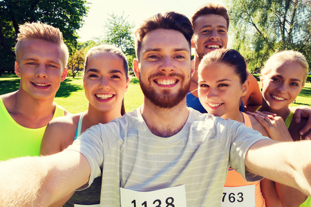 fitness, sport, friendship, technology and healthy lifestyle concept - group of happy teenage friends with racing badge numbers taking selfie by smartphone at marathon outdoorsの写真素材