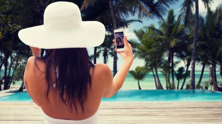 travel, tourism, summer, technology and people concept - smiling young woman or teenage girl in sun hat taking selfie with smartphone over resort beach with palms and swimming pool backgroundの写真素材