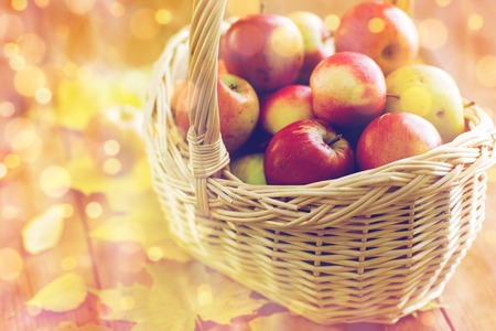 gardening, season, autumn and fruits concept - close up of wicker basket with ripe red apples and leaves on wooden tableの写真素材