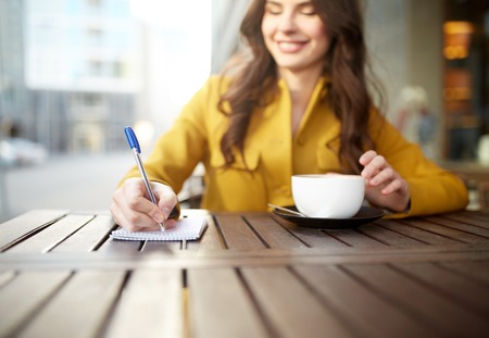 leisure and people concept - happy young woman or teenage girl with cup drinking cappucino and writing to notebook at city street cafe terraceの写真素材