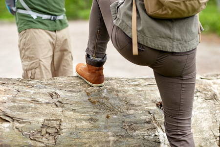 adventure, travel, tourism, hike and people concept - close up of couple with backpacks walking and climbing over fallen tree trunk in woodsの写真素材
