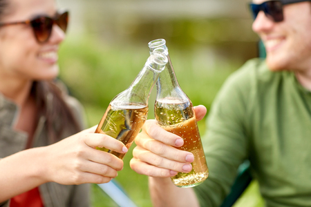travel, hike, drinks, leisure and people concept - close up of happy couple clinking glass bottles and drinking cider or beer at campsiteの写真素材